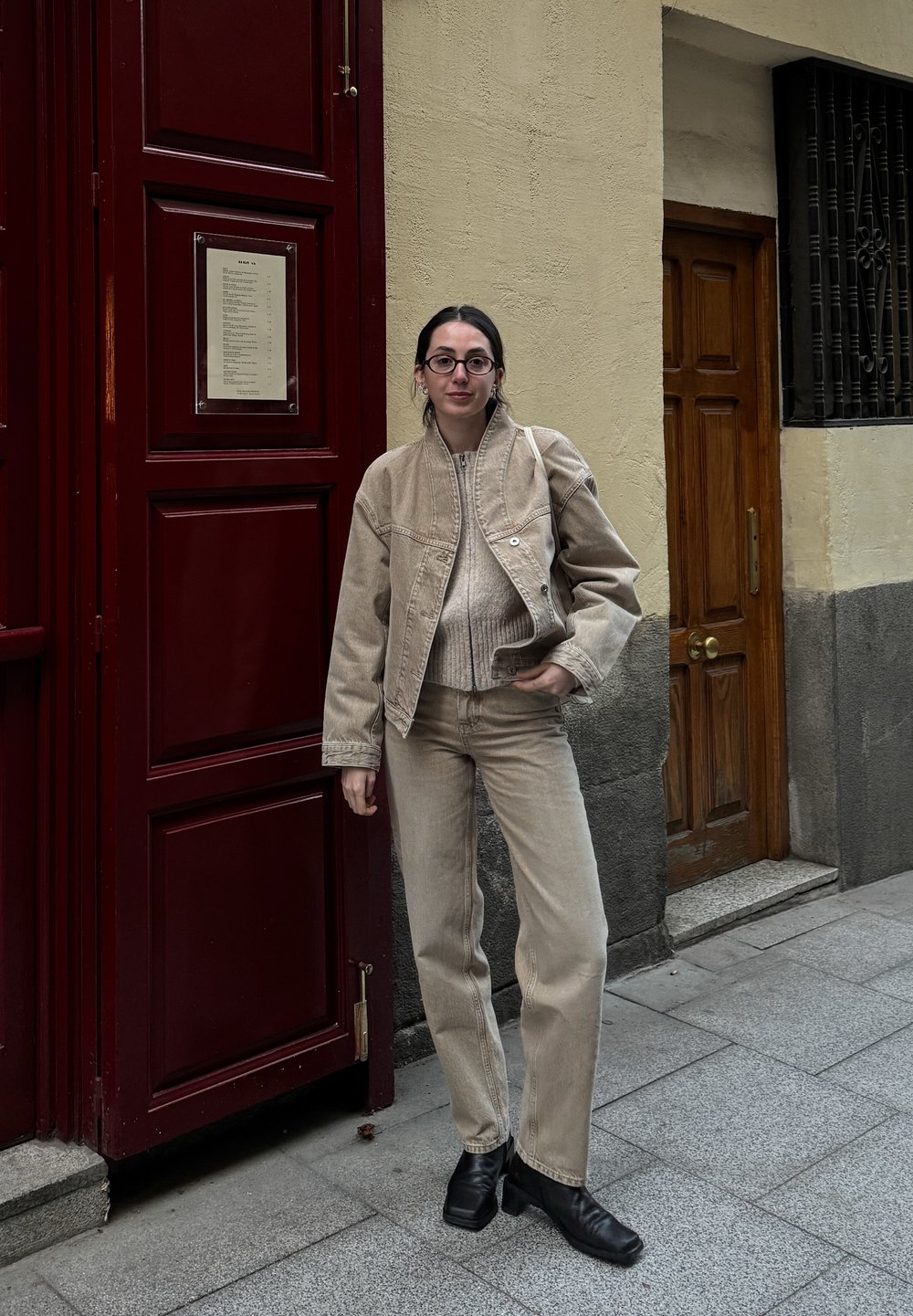 Beige kordbársony zakó és passzoló nadrág, fekete bőr bokacsizmával párosítva. A háttérben vörös ajtó és világos fal található.