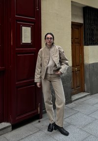Beige kordbársony zakó és passzoló nadrág, fekete bőr bokacsizmával párosítva. A háttérben vörös ajtó és világos fal található.