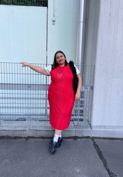 Vestido midi rojo acanalado con mangas cortas, combinado con un chaleco de piel negra. Zapatos gruesos negros usados con calcetas blancas. Collar con colgante en forma de corazón plateado.