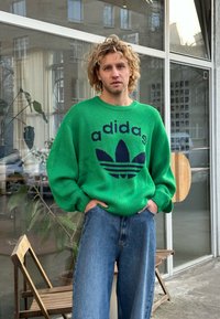 Πράσινη πλεκτή ζακέτα με μπλε λογότυπο "adidas" και σχέδιο τριφύλλι, συνδυασμένη με φαρδιά τζιν παντελόνια, σε έναν αστικό εσωτερικό χώρο.