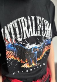 Chemise noire avec un grand texte blanc fissuré "NATURAL" et un aigle coloré aux ailes bleues et orange déployées, texte "NEVERSL" en dessous.