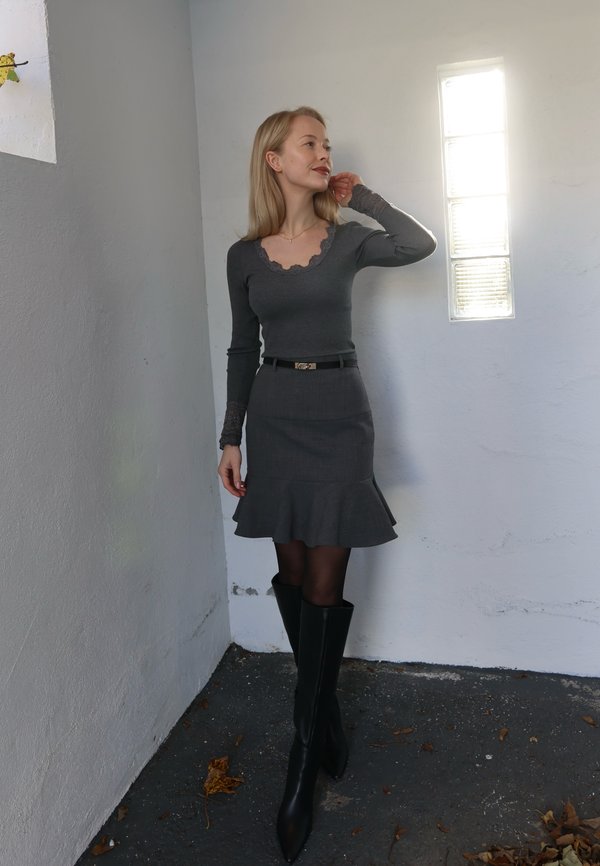 Top de manga larga gris con escote de encaje, combinado con una falda gris de vuelo y botas negras hasta el muslo, de pie contra una pared blanca.