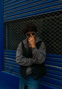 Μαύρο φουσκωτό γιλέκο πάνω από γκρίζα φούτερ, μπλε τζιν και γυαλιά ηλίου. Σηκωμένος μπροστά σε γαλάζια μεταλλική πόρτα με υπόβαθρο από εξαγωνικό μοτίβο.