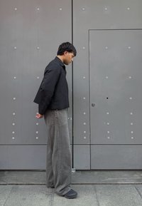 Čierna, priliehavá bunda cez košeľu s vysokým golierom, spárovaná s širokými sivými džínsy a čiernymi topánkami; zasadená oproti šedej stene s kruhovými študentmi.