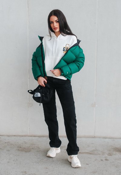 Chaqueta de plumas verde sobre una sudadera blanca con gráfico, pantalones de pana negros y zapatillas blancas. Incluye una pequeña bolsa negra con un logo.