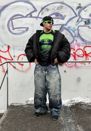 Hombre con vaqueros anchos, chaqueta acolchada negra, camiseta verde y negra de DC, gorra negra y gafas de sol verdes de pie junto a una pared con grafitis.
