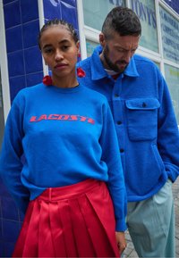 Sweatshirt azul com o logótipo "LACOSTE" vermelho, combinado com uma saia plissada vermelha. Casaco de fleece azul no homem, calças azul claro. Brincos florais vermelhos.