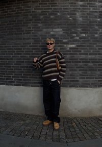 Pull en laine rayé aux tons bruns, crème et foncés, associé à un jean sombre et des chaussures marron clair. Accessoires : lunettes de soleil et sac marron.