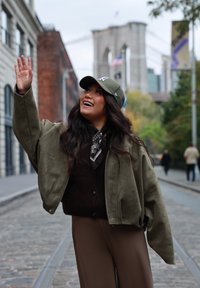 Veste bomber vert olive, pull en maille foncé, pantalon marron, écharpe à motifs noir et blanc, casquette grise, arrière-plan urbain avec un mur en briques.