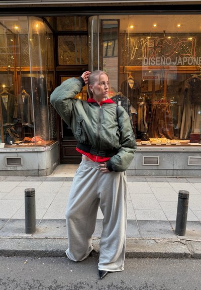 Joven con una chaqueta bomber verde de gran tamaño y pantalones anchos grises posa en la acera de la ciudad frente a la vitrina de una tienda de ropa.