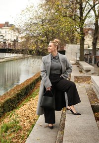 Szary płaszcz, koszula na guziki, czarne spodnie o szerokich nogawkach i czarne szpilki. Trzyma czarną torebkę, w tle rzeka i jesienne liście.