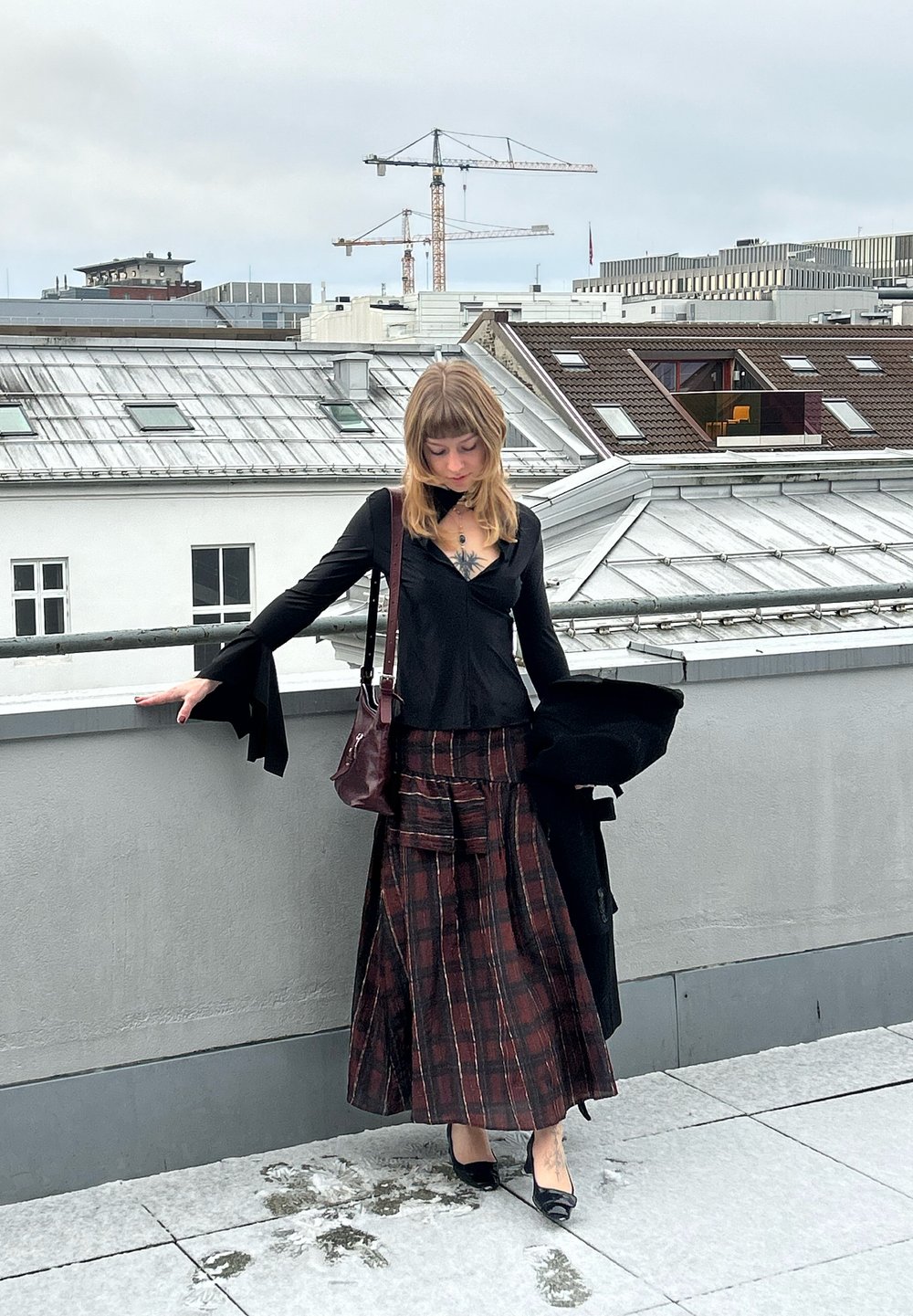 Bluză neagră cu mâneci lungi și manșete evazate, asortată cu o fustă lungă în carouri roșu închis și negru. Poartă o geantă burgundy și pantofi cu toc negri.