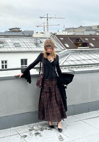 Bluză neagră cu mâneci lungi și manșete evazate, asortată cu o fustă lungă în carouri roșu închis și negru. Poartă o geantă burgundy și pantofi cu toc negri.