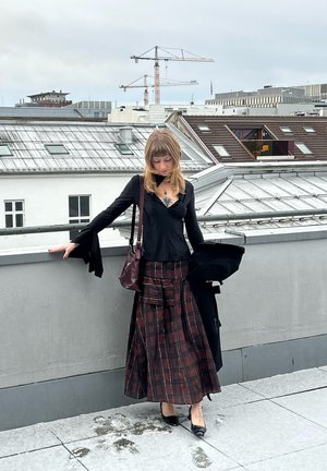 Top negro de manga larga con puños acampanados, combinado con una falda larga de cuadros en rojo oscuro y negro. Lleva un bolso burdeos y tacones negros.