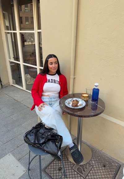 Joven mujer con cárdigan rojo y camiseta blanca con la palabra "Madrid" sentada en una mesa de café al aire libre con galletas, café y una botella, junto a una pared beige.