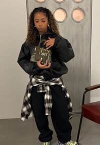Jeune femme en veste noire et chemise à carreaux nouée à la taille, tenant un petit sac à main à motifs, debout près d'une porte en métal et d'une chaise rouge.