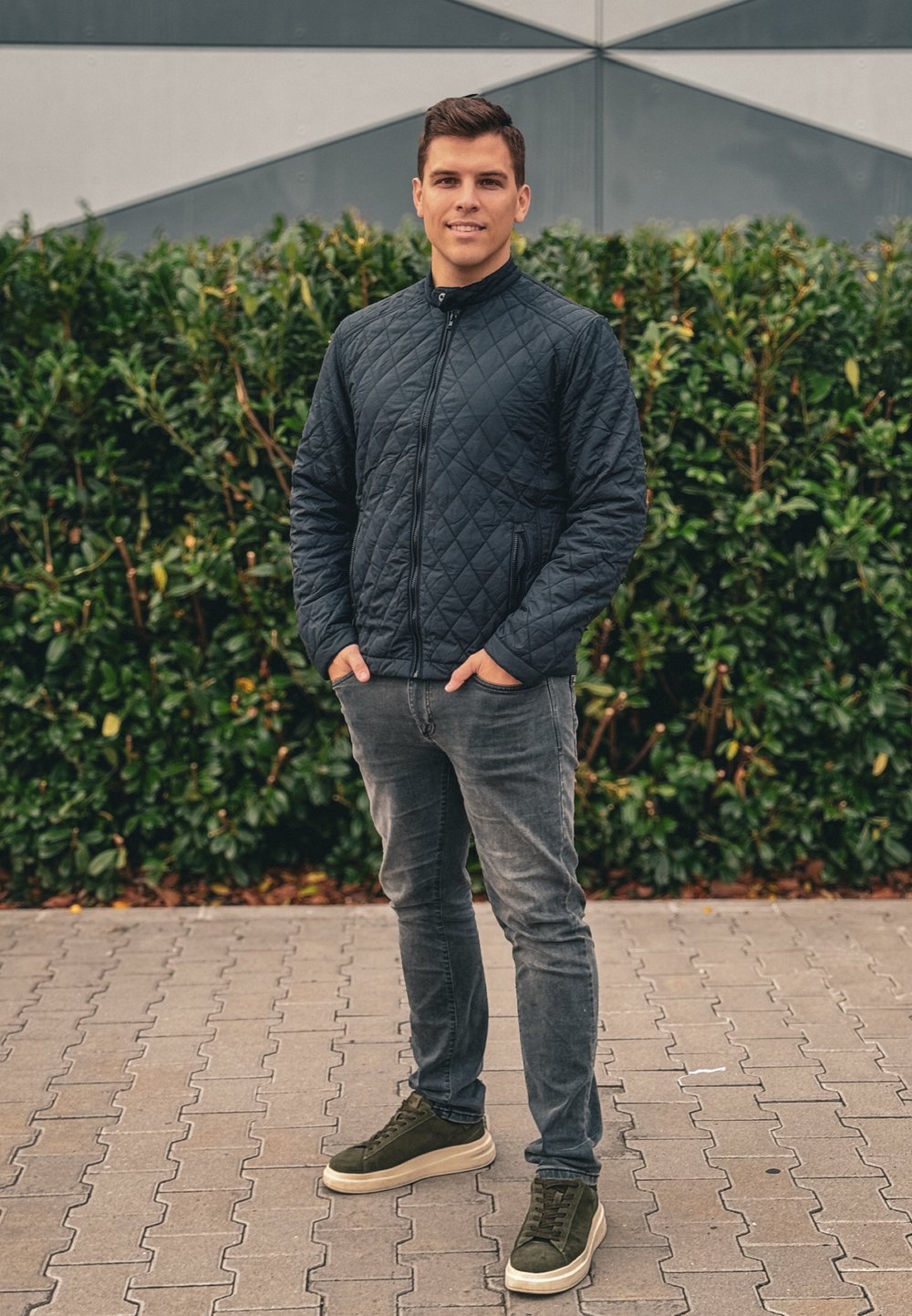 Πετσέτα μαύρης ραφής, γκρι τζιν και πράσινα αθλητικά. Η πετσέτα διαθέτει ψηλό γιακά και φερμουάρ μπροστά. Φόντο από πράσινη βλάστηση και γεωμετρικό τοίχο.