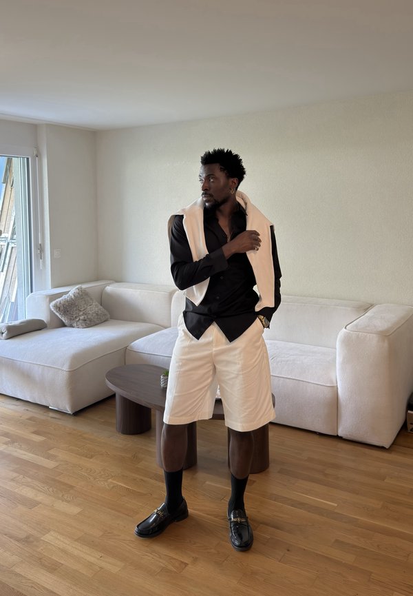 Mand står i stuen iført sort skjorte, beige shorts, beige sweater over skuldrene, sorte sokker og sorte loafers.
