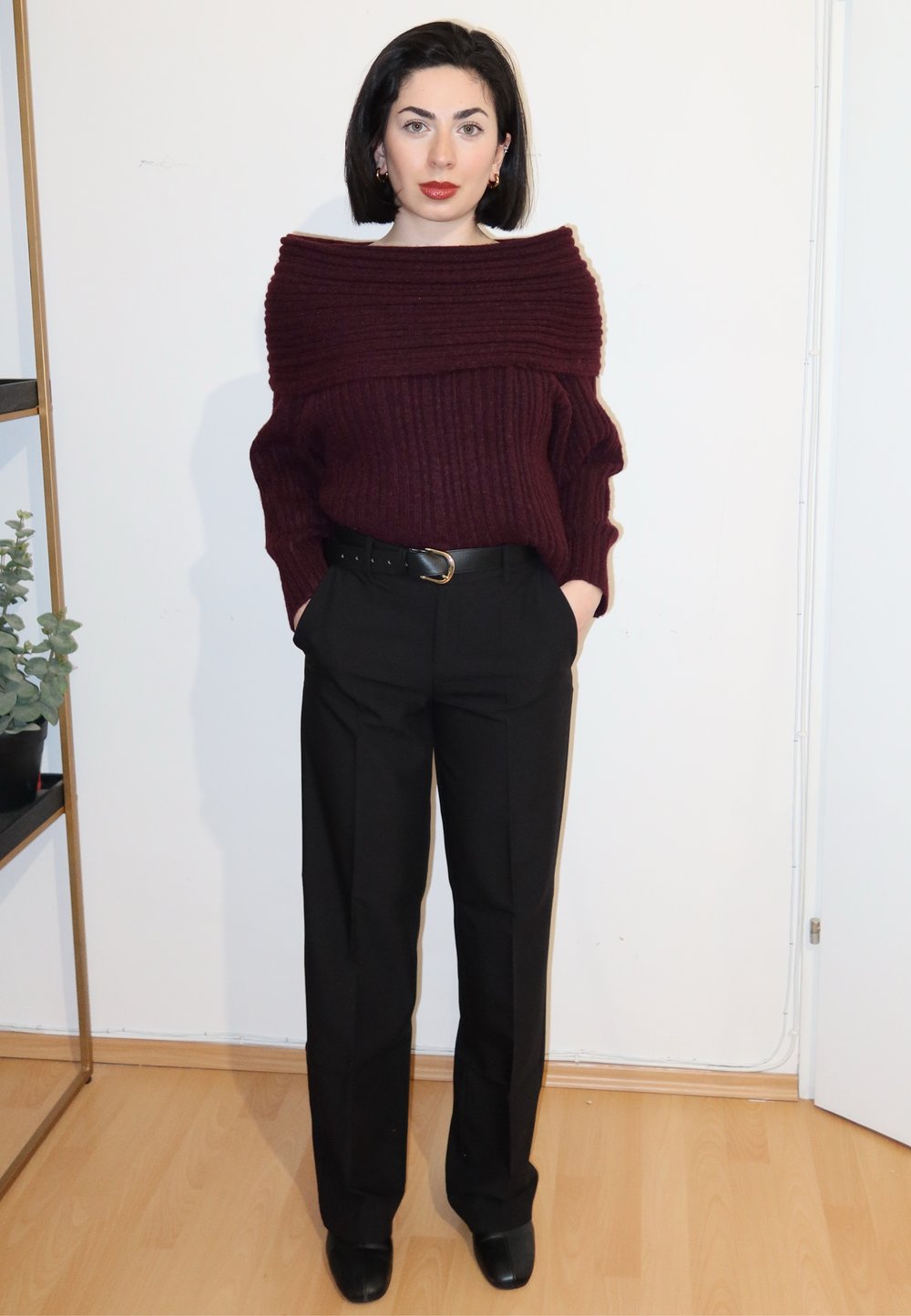 Femme aux cheveux courts et foncés portant un pull bordeaux à épaules dénudées, un pantalon noir avec ceinture et des chaussures noires, debout à l'intérieur.