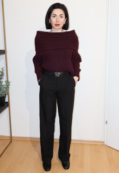 Mujer con cabello corto y oscuro, vistiendo un suéter burdeos sin hombros, pantalones negros con cinturón y zapatos negros, de pie en el interior.