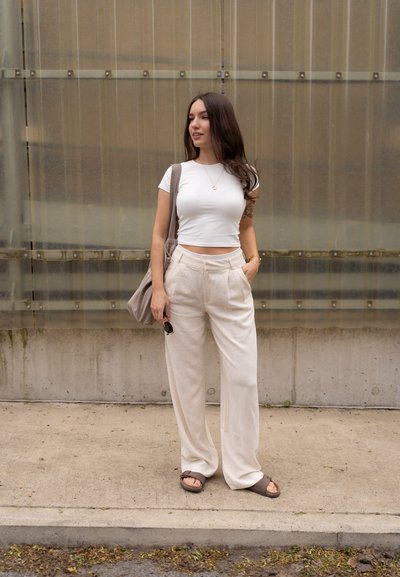 Camiseta blanca corta y pantalones de lino beige de pierna ancha. Lleva un bolso gris al hombro y sandalias marrones. El fondo presenta paneles de vidrio verticales.