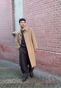 Manteau en laine beige porté sur un pull sombre, pantalon gris large et bottes noires à lacets. Fond mural en briques avec pavé texturé.