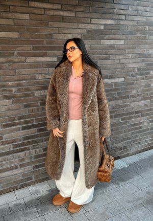 Abrigo de piel sintética marrón con un collar ancho, camiseta rosa claro, pantalones color crema y zapatos de plataforma marrones, complementados con un pequeño bolso marrón.