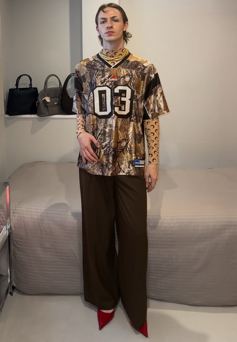 Jeune personne avec un maillot Adidas camouflage par-dessus un haut à manches longues à motifs, un pantalon large marron et des talons aiguilles rouges, debout dans une chambre.