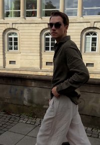 Alyvuogių žalios spalvos marškiniai su mygtukais ir šviesiai smėlinio lino kelnės, stovinčios prieš akmeninį pastatą su aukštais langais ir pilku akmeniniu taku.