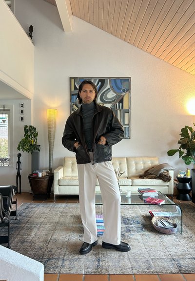 Chaqueta de cuero negro con cuello alto, suéter gris de cuello vuelto, pantalones blancos y zapatos negros con plataforma, sobre un fondo de una sala de estar moderna.