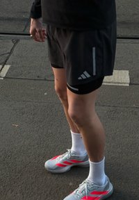 Személy fekete Adidas rövidnadrágban, fehér bokazokniban és szürke, piros csíkkal díszített sportcipőben áll az aszfalton.