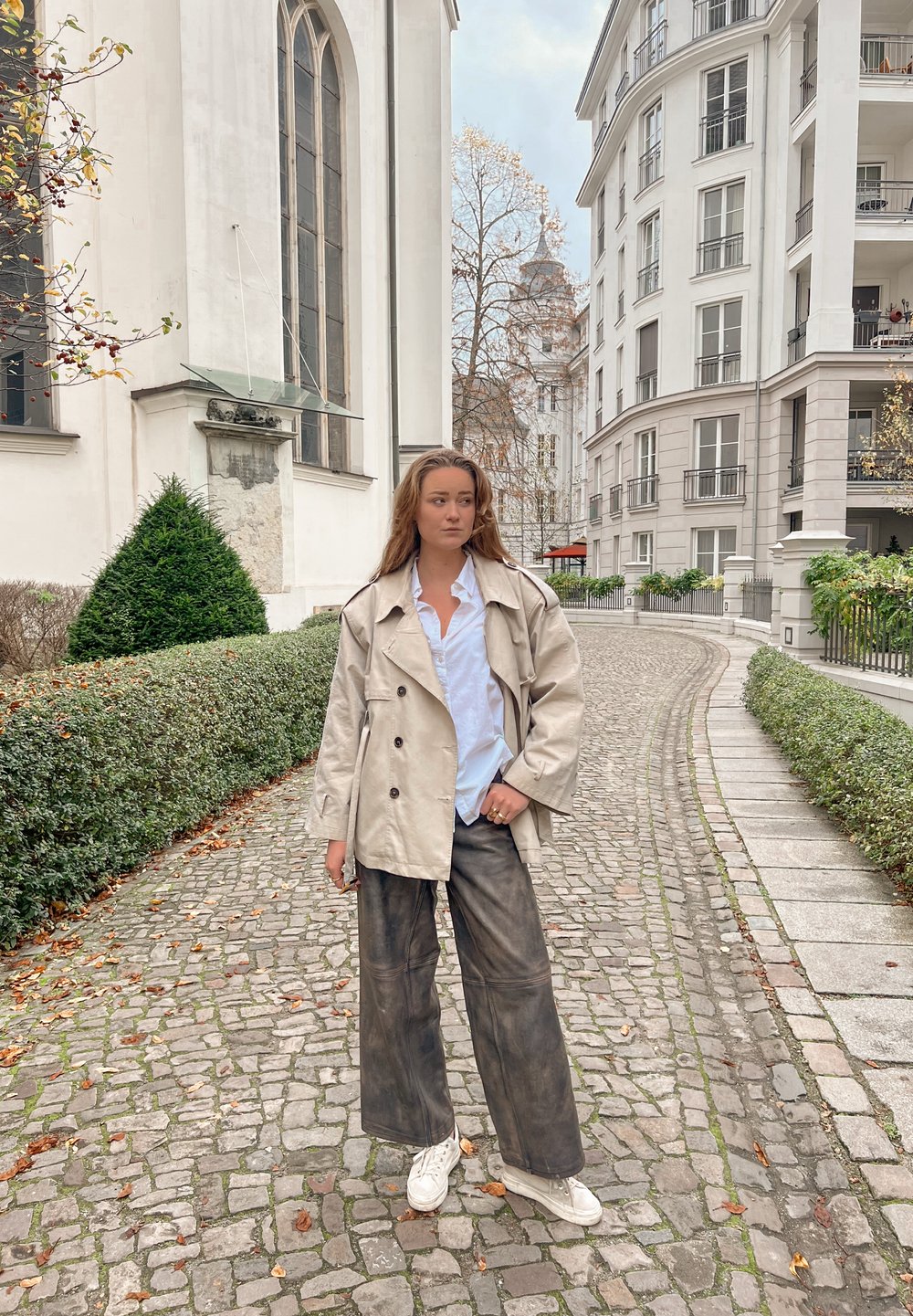 Trench-coat beige croisé porté sur une chemise blanche, associé à un pantalon large noir et des baskets blanches ; posé sur un chemin pavé avec de la verdure.