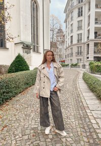 Trench-coat beige croisé porté sur une chemise blanche, associé à un pantalon large noir et des baskets blanches ; posé sur un chemin pavé avec de la verdure.