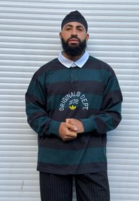 Homme avec barbe portant un maillot de rugby rayé noir et vert avec un col blanc et un pantalon à fines rayures, debout devant un rideau métallique.