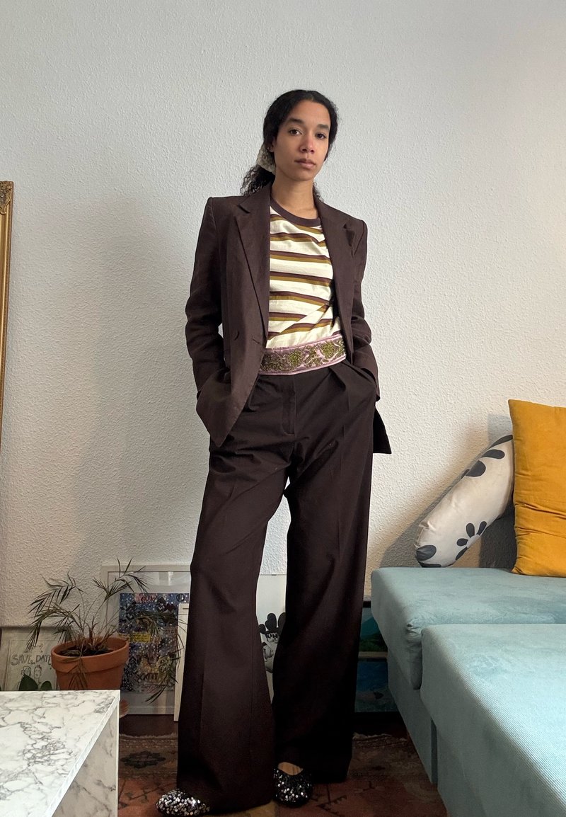 Femme debout à l'intérieur, portant un costume marron, une chemise rayée et une ceinture à motifs, à côté d'un canapé bleu sarcelle avec des coussins jaunes et à motifs.