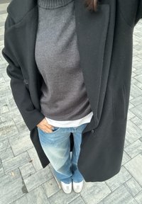 Oseba, oblečena v črni plašč, siv pulover, belo srajco, modre kavbojke in bele superge, stoji na sivem tlaku z eno roko v žepu.