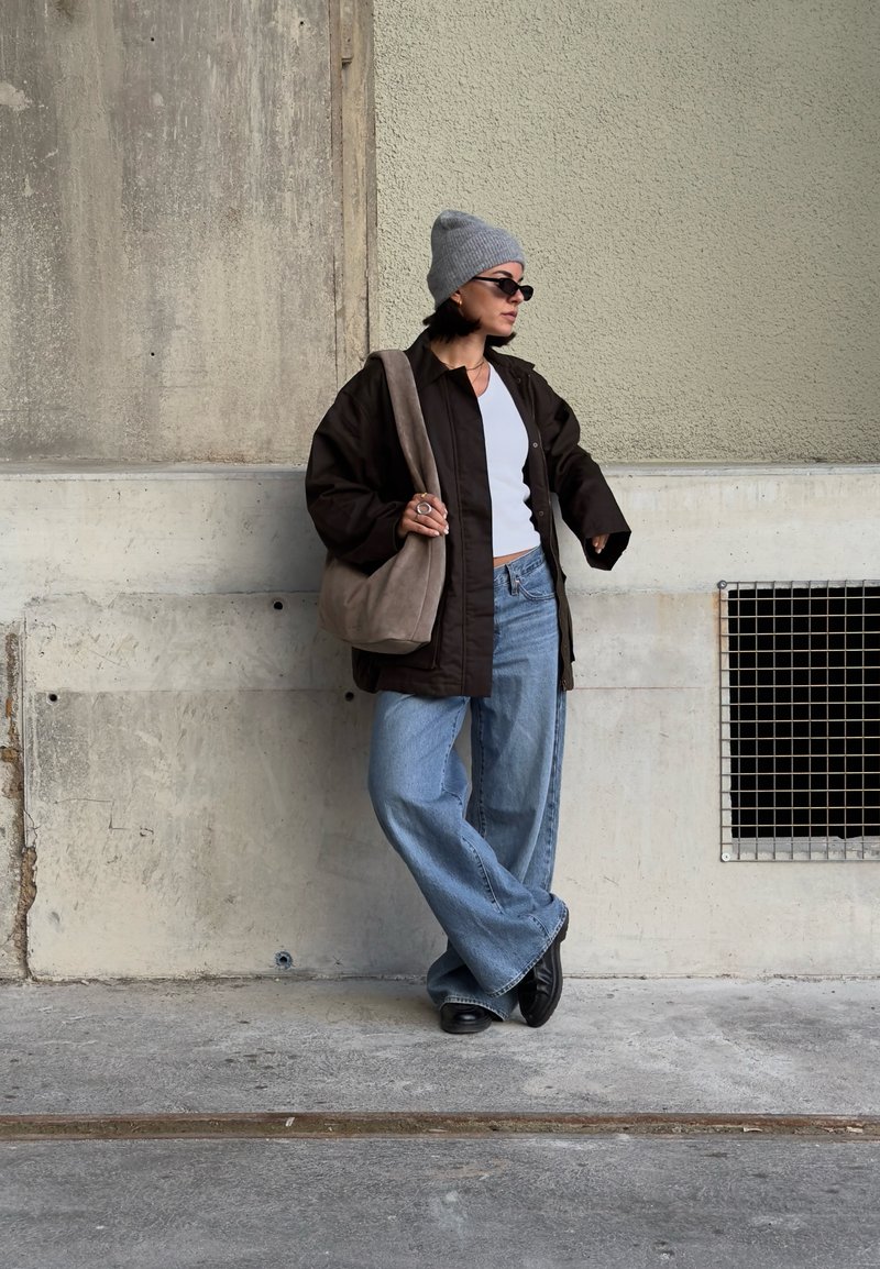 Casaco oversized castanho, top branco de alças, jeans de perna larga azul claro, gorro cinzento e sapatos pretos. Carrega uma mala de ombro cinzenta. Fundo texturado na parede.