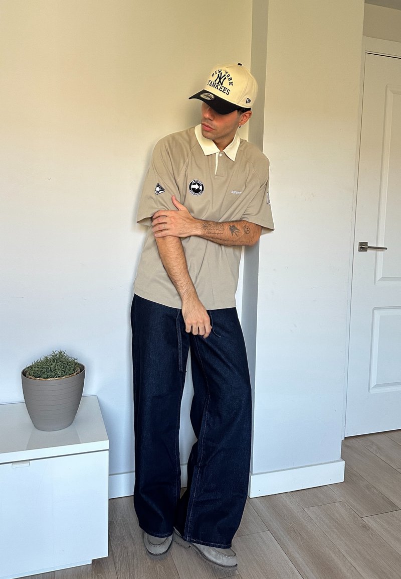 Polo beige avec un col blanc et des accents de logo, associé à un jean bleu foncé ample et une casquette de baseball beige avec du texte noir.