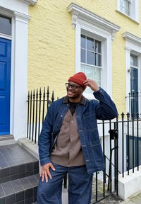 Jachetă din denim peste un pulover maro tricotat, asortată cu pantaloni largi de aceeași culoare. Căciulă tricotată roșie și ochelari completează ținuta. Fundal cu perete de cărămidă galbenă.