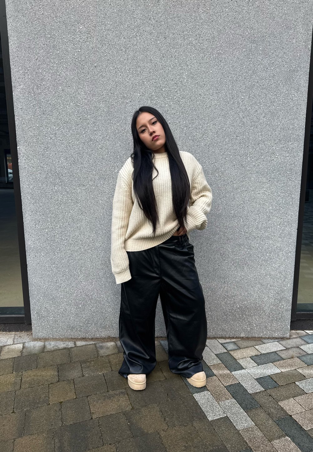 Pullover oversized in maglia beige, pantaloni wide-leg in finta pelle nera e scarpe platform beige. In piedi contro un muro grigio strutturato.