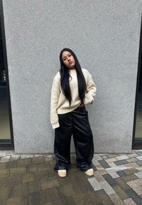 Pullover oversized in maglia beige, pantaloni wide-leg in finta pelle nera e scarpe platform beige. In piedi contro un muro grigio strutturato.