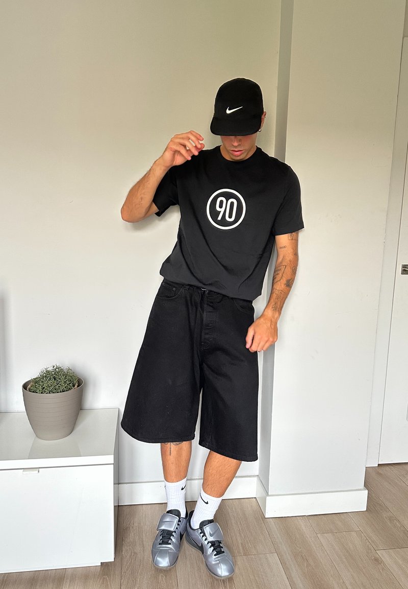T-shirt noir à manches courtes avec un motif circulaire blanc portant "90." Shorts en denim noir, chaussures de sport argentées et chaussettes blanches Nike.