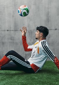 Weißes Fußballtrikot mit roten und schwarzen Akzenten, schwarze Hose mit weißen Streifen, sitzt auf Kunstrasen und jongliert mit einem mehrfarbigen Fußball.