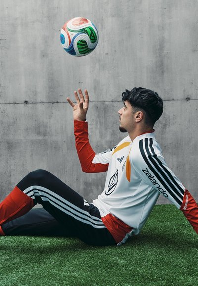 Camiseta de fútbol blanca con acentos rojos y negros, pantalones negros con rayas blancas, sentado sobre césped sintético, haciendo malabares con un balón de fútbol multicolor.