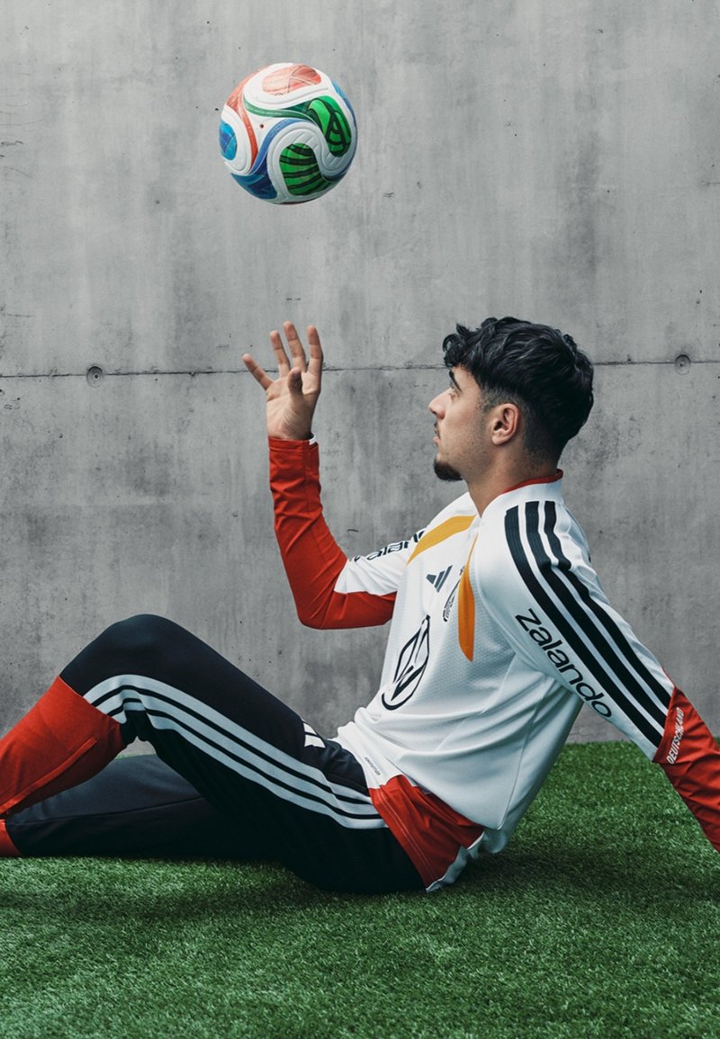 Maillot de soccer blanc avec des accents rouges et noirs, pantalons noirs avec des rayures blanches, assis sur de l'herbe synthétique, jonglant avec un ballon de soccer multicolore.