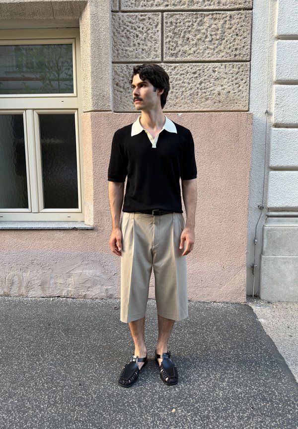 Hombre con cabello oscuro y bigote, vestido con una camisa negra con cuello blanco, pantalones cortos beige hasta la rodilla y sandalias negras, de pie en una acera de la ciudad.