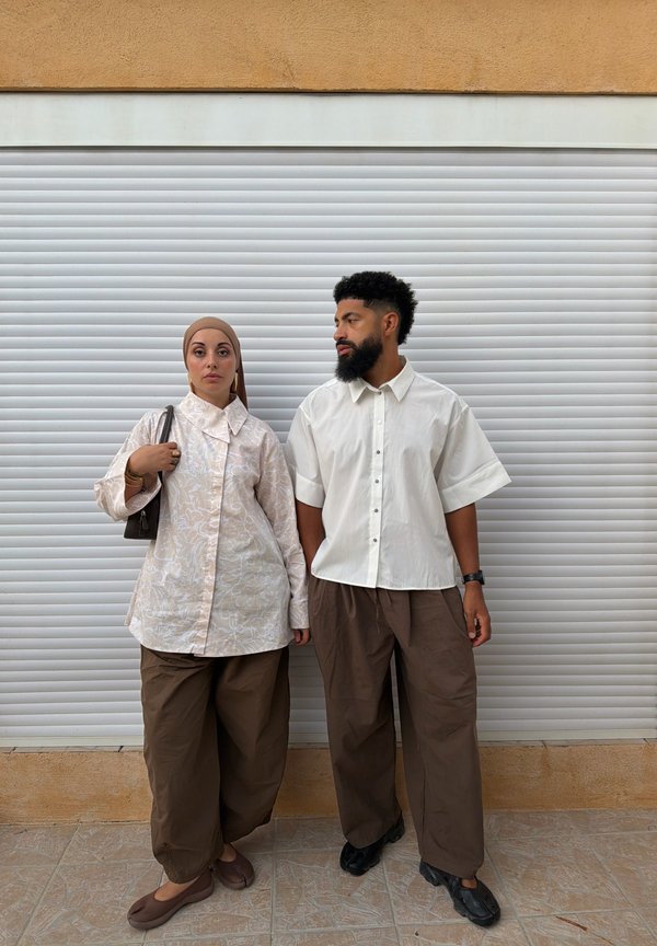 Una mujer con un pañuelo en la cabeza y un hombre con barba están uno al lado del otro frente a una persiana blanca, ambos vestidos con pantalones marrones holgados y camisetas claras.