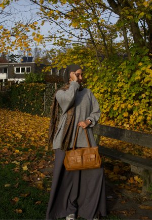 Bolso de cuero marrón con dos bolsillos frontales, suéter gris oversized y falda larga gris, sobre un fondo de hojas otoñales.