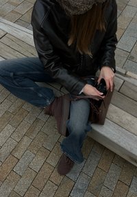 Personne portant un chapeau en fourrure, une veste en cuir noire et un jean est assise sur un banc en bois, rangeant un appareil photo dans un sac en cuir marron.