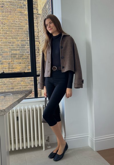 Chaqueta corta de cuadros marrones con botones sobre un jersey negro y pantalones negros ajustados, combinada con zapatos planos negros y un bolso pequeño.